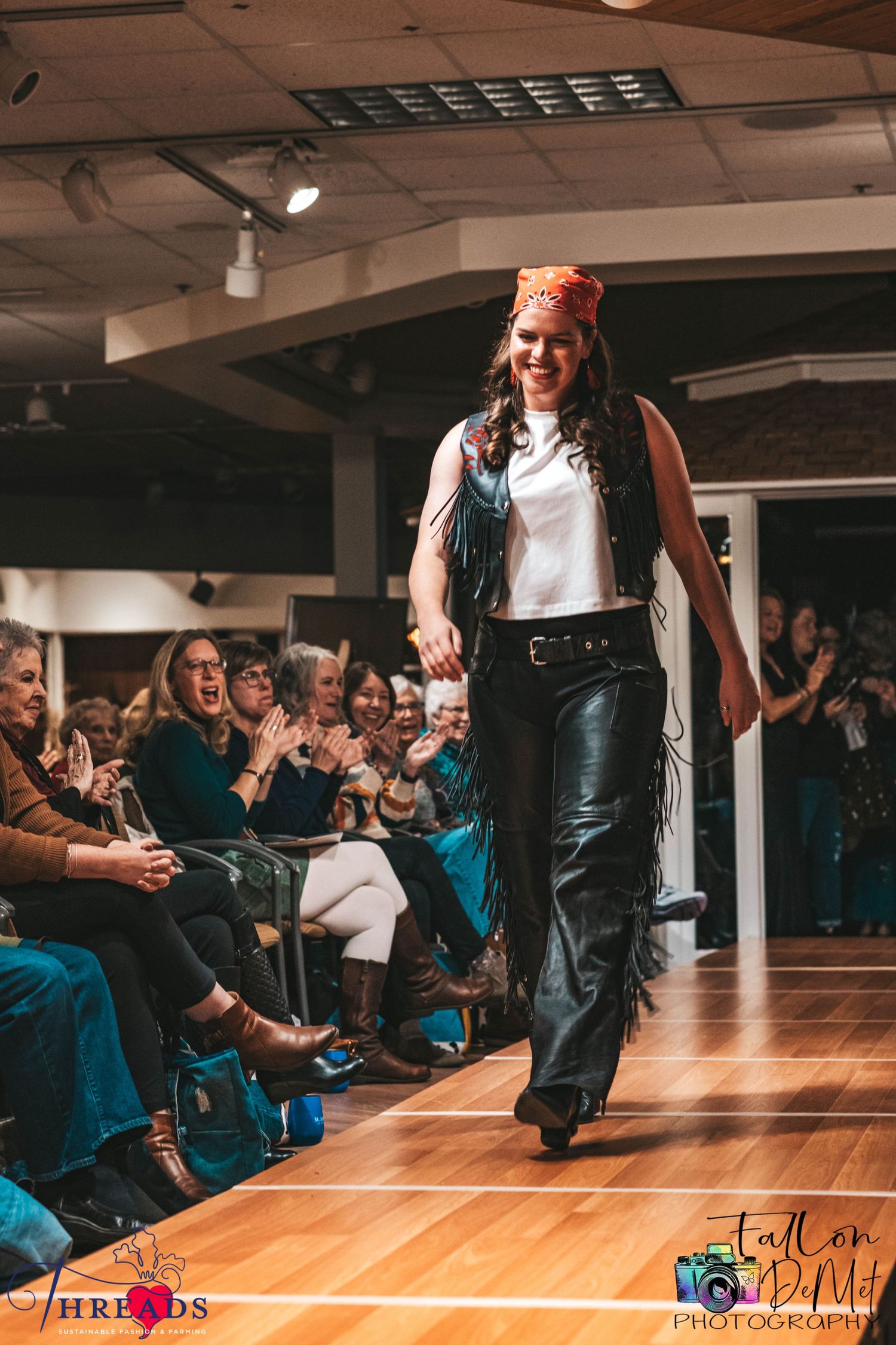 Fashion model walking runway in leather pants and long vest while audience watches.