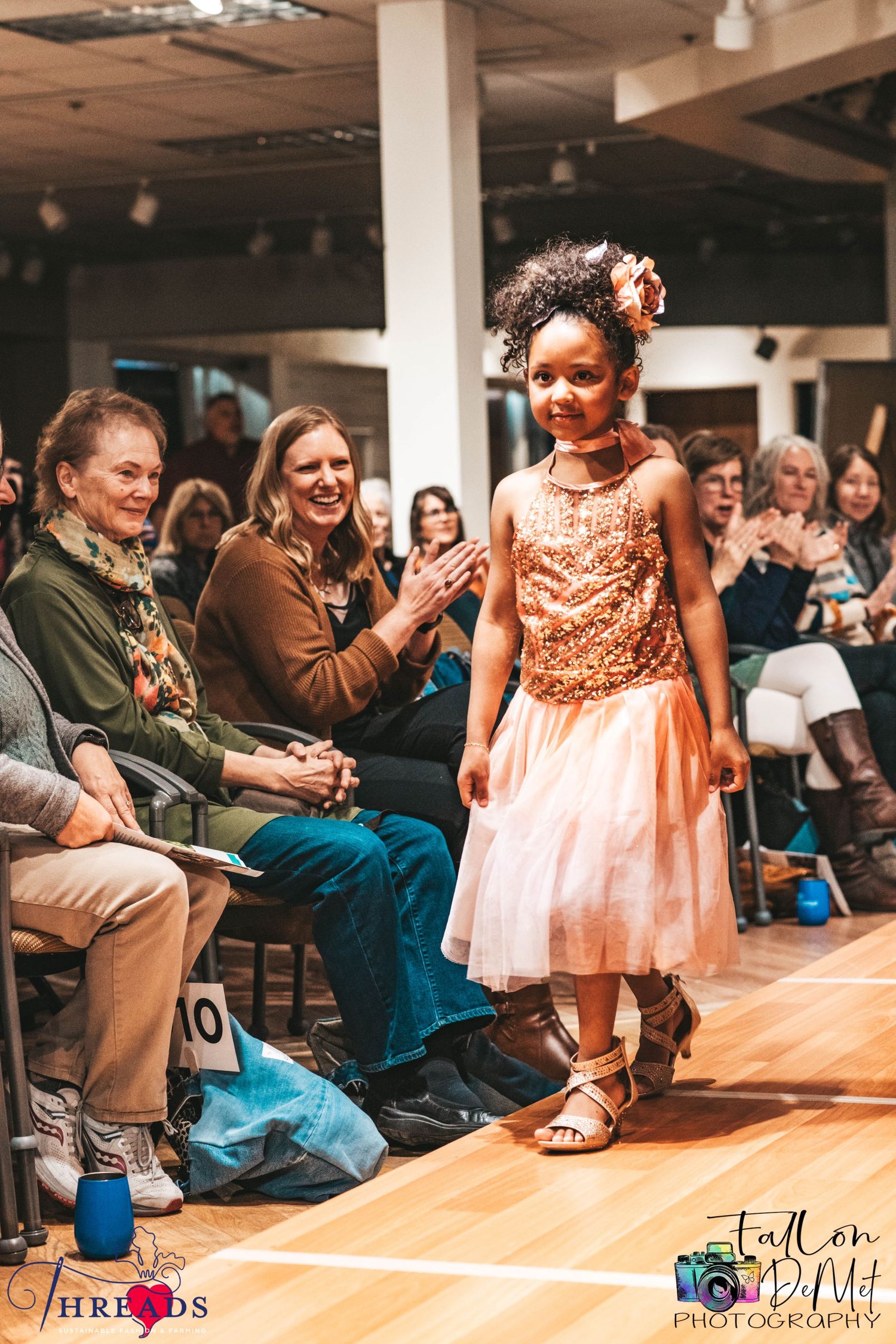 Little girl in peach tulle dress walking runway as audience applauds at fashion show.
