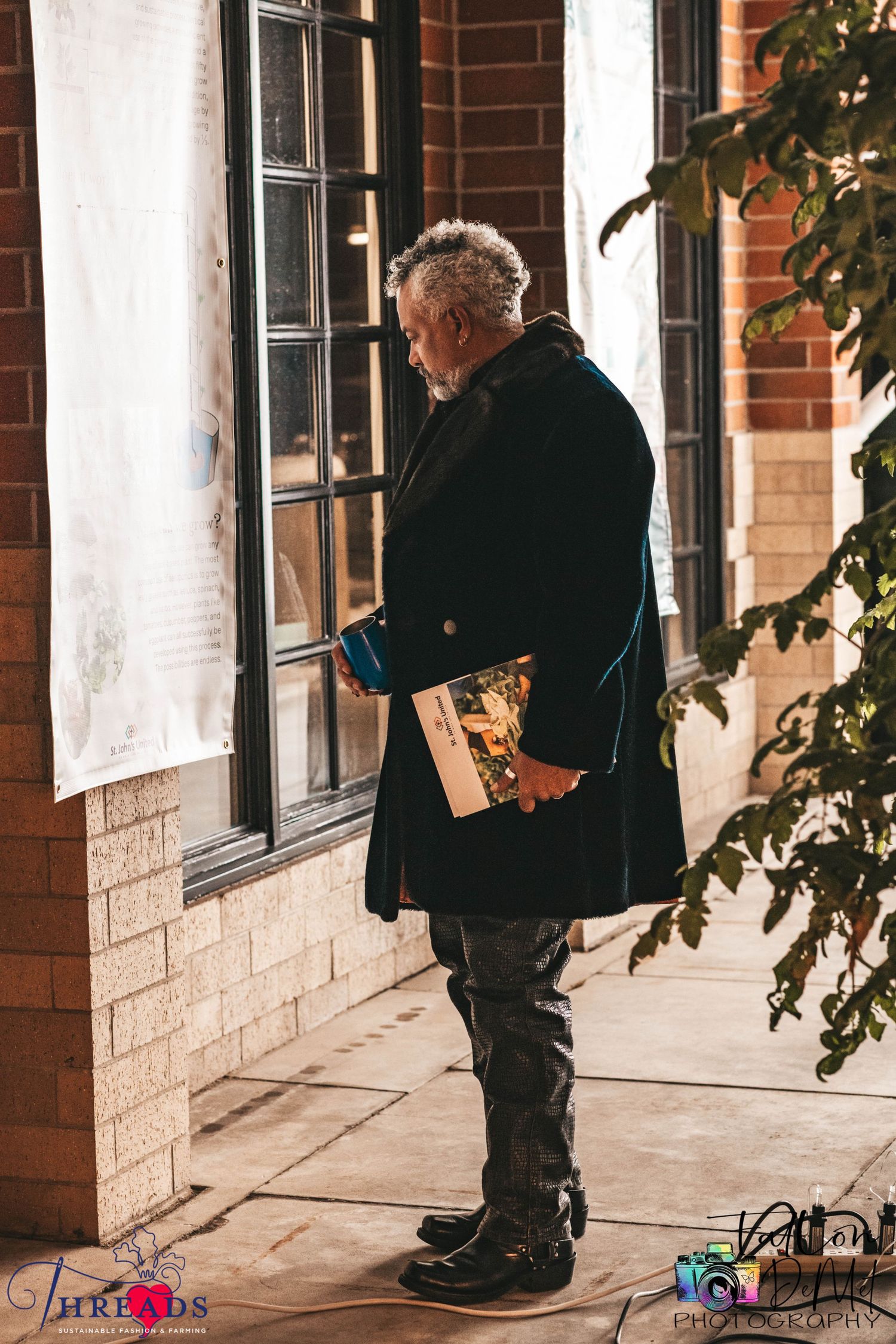 An elderly figure in a black coat carries a book while standing on a brick building's porch.