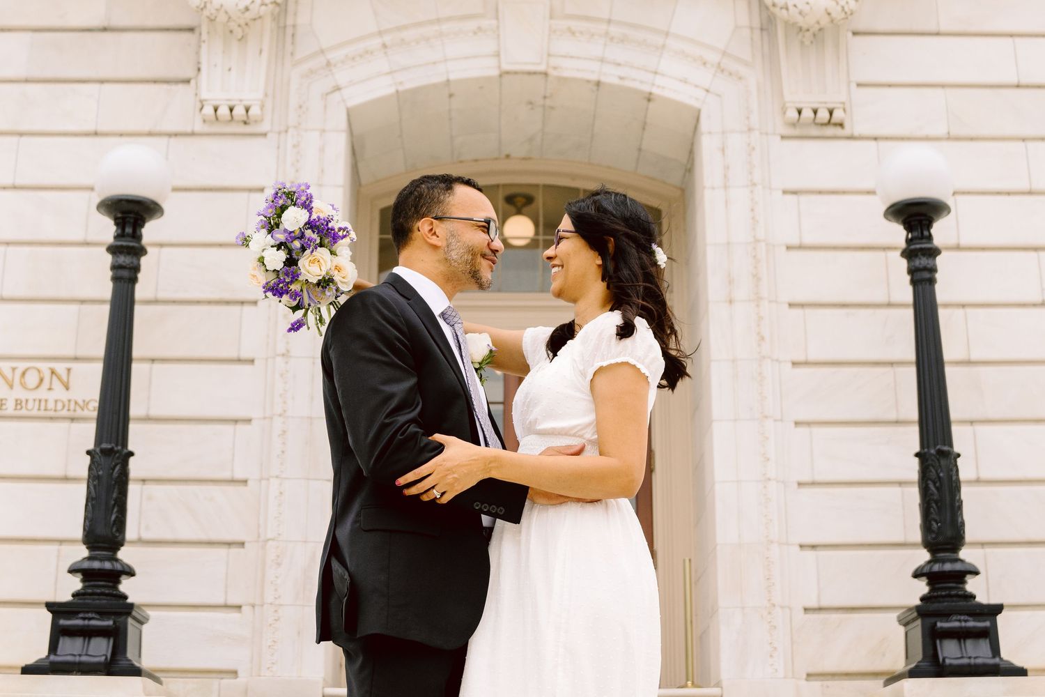 A romantic wedding couple shares a tender moment against a classic white building exterior with ornate columns.