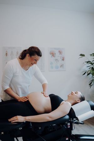 Eine Chiropraktorin behandelt eine Schwangere im Behanldungsraum. Die Schwangere liegt dabei auf dem Tisch und lacht die Chriopraktorin an.