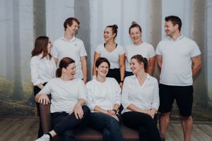 Ein Chiropraktorenteam guckt sich strahelnd an, in einem Sportraum vor einer Waldtapete. Jeder trägt ein weißes T-Shirt und lacht.