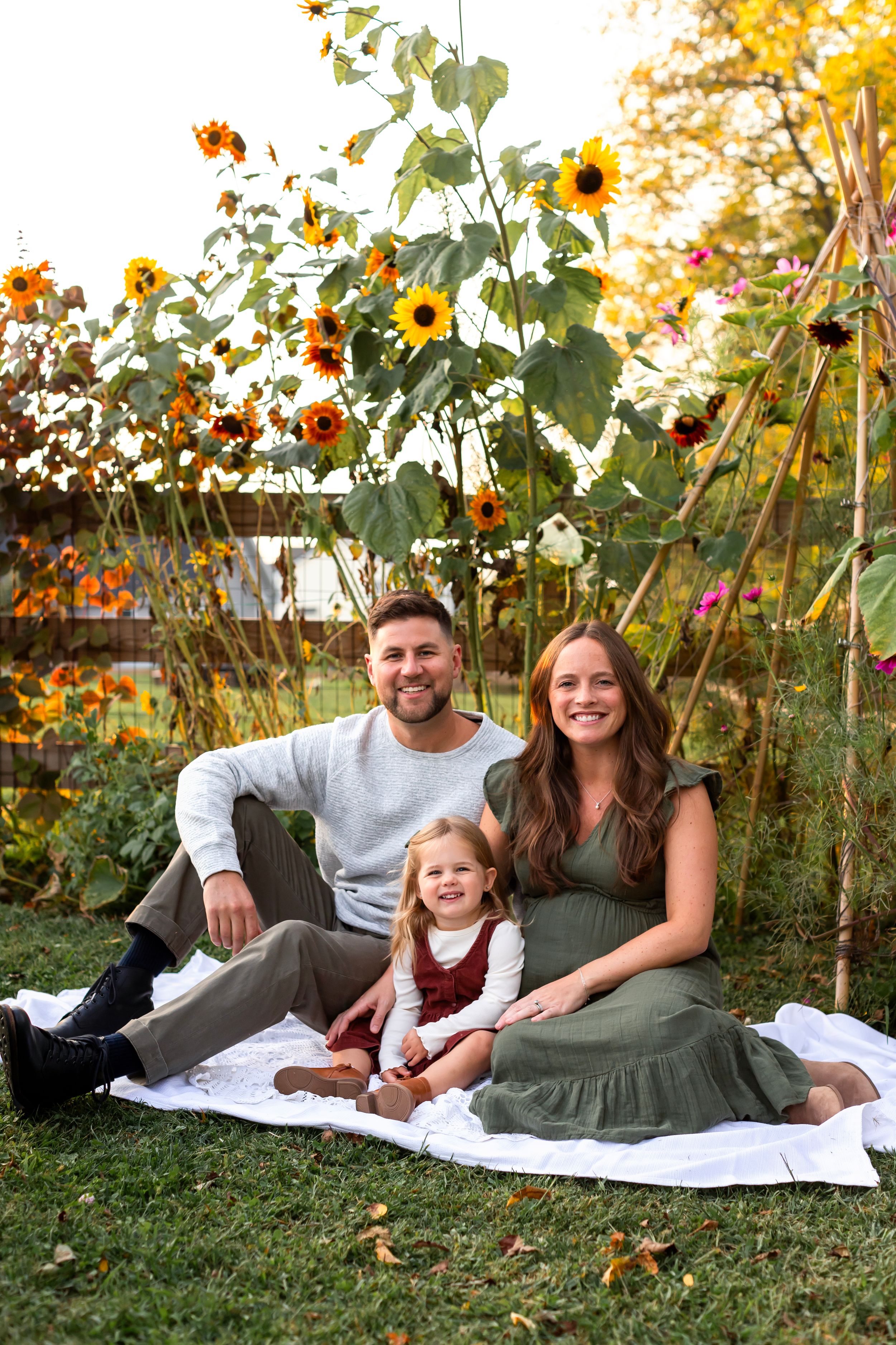 Family sitting on white blanket in garden with tall sunflowers and autumn foliage.
