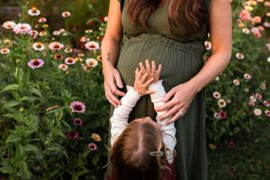 Small hands reach toward a maternity dress in a garden filled with pink flowers.
