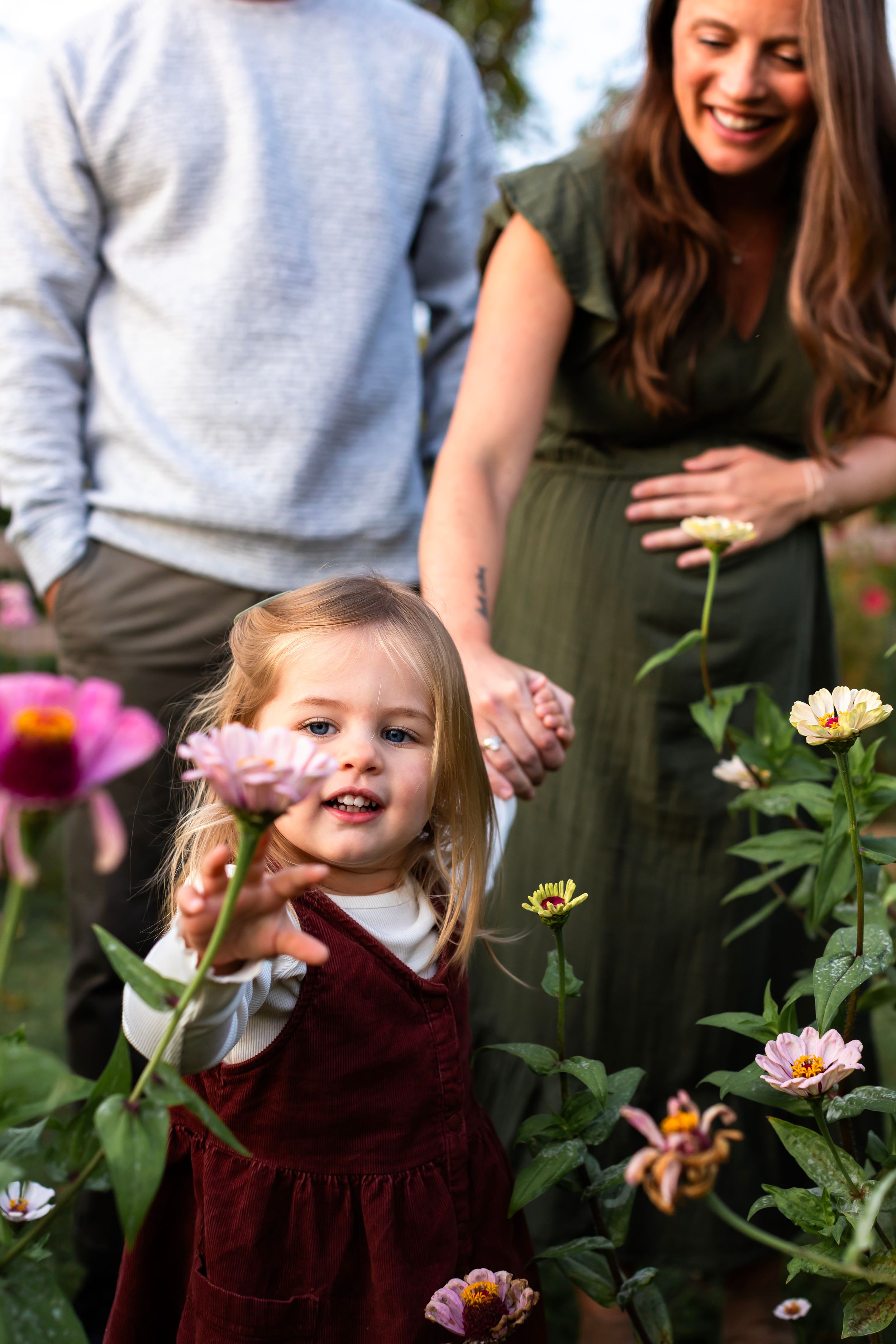 Child in burgundy dress reaching for flowers in garden while parents watch.