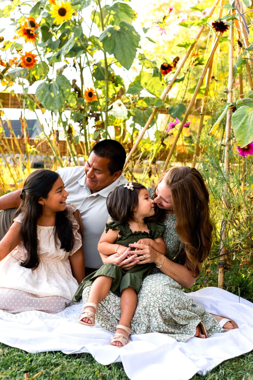 Family relaxing on white blanket in garden with blooming sunflowers and summer flowers.
