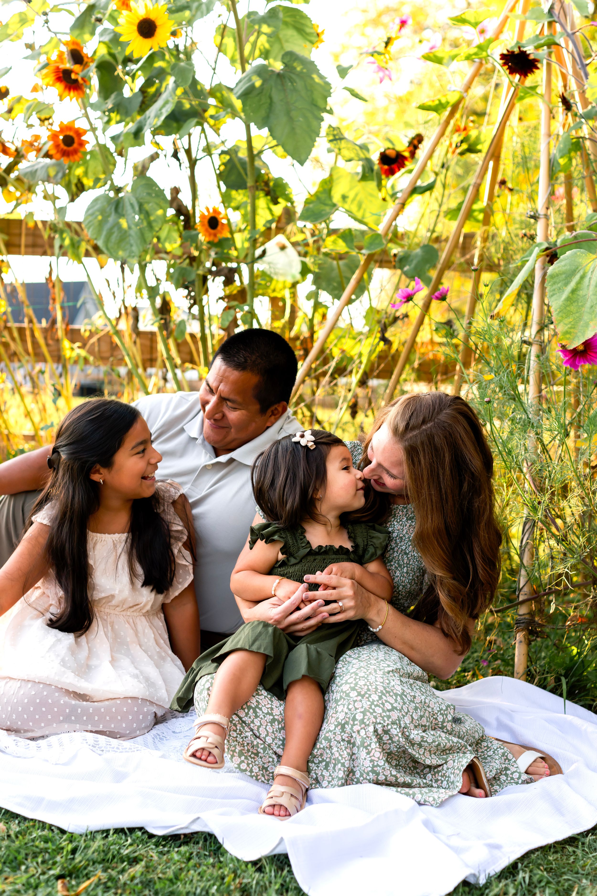 Family relaxing on white blanket in garden with blooming sunflowers and summer flowers.