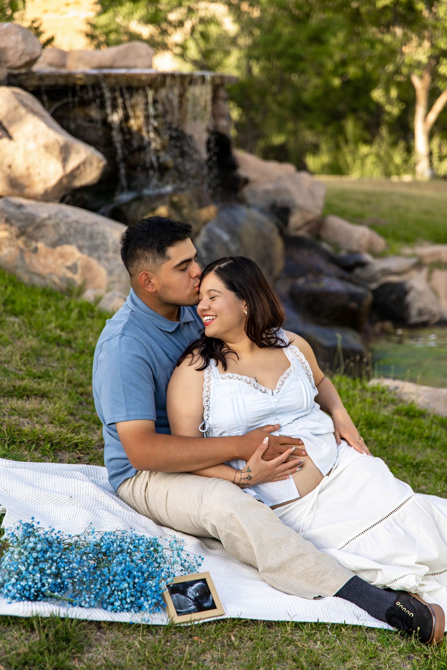 A couple sitting on a picnic blanket by a waterfall garden, with blue flowers and an ultrasound photo visible.
