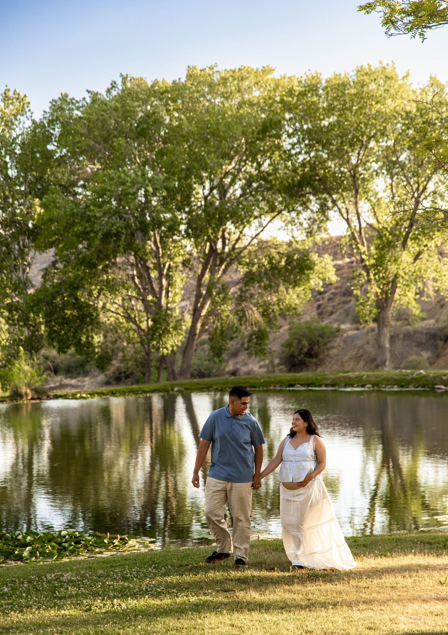 A couple walks hand in hand along a serene pond surrounded by lush trees at sunset.