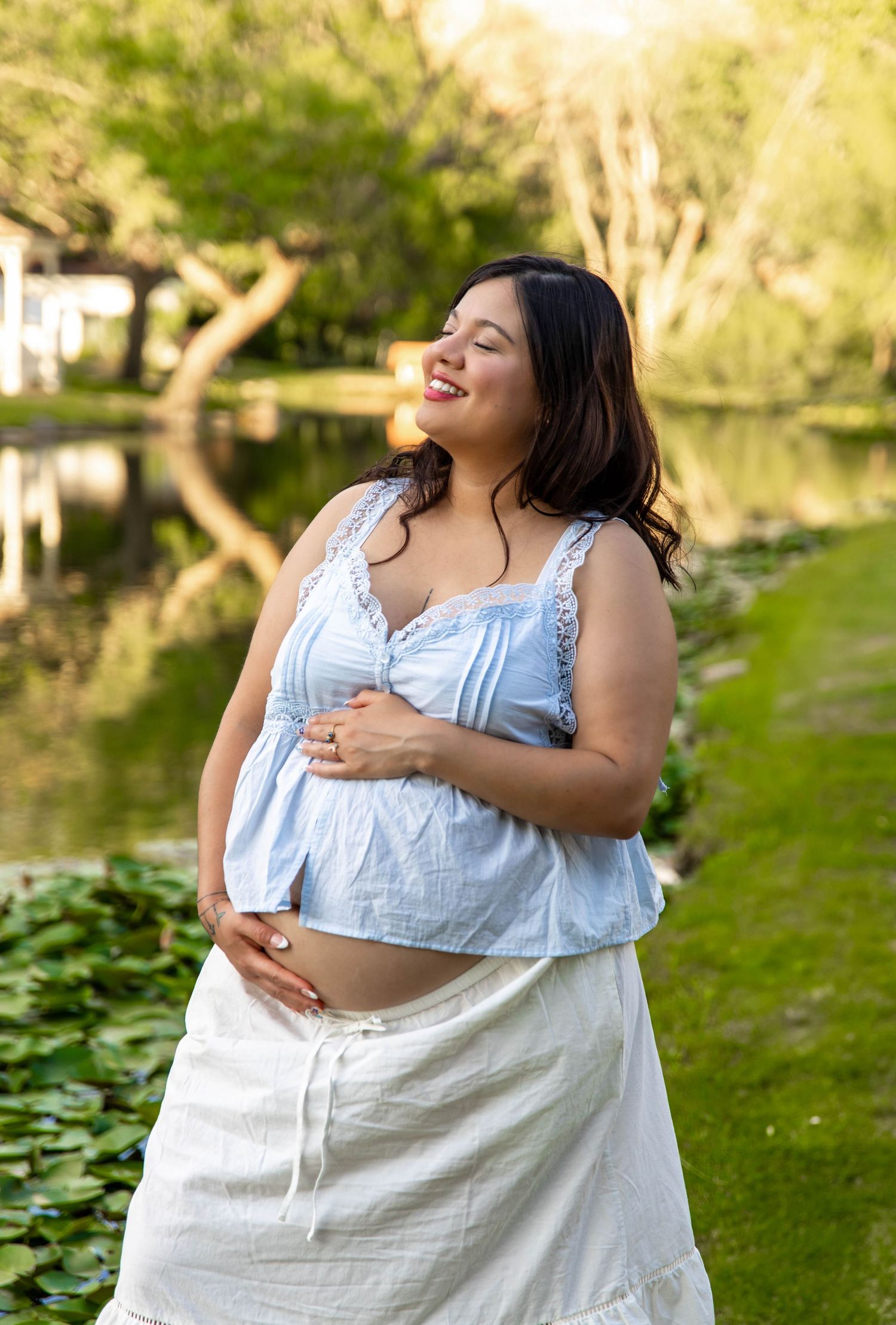 A woman in a white dress laughs joyfully while sitting outdoors near a pond on a sunny day.
