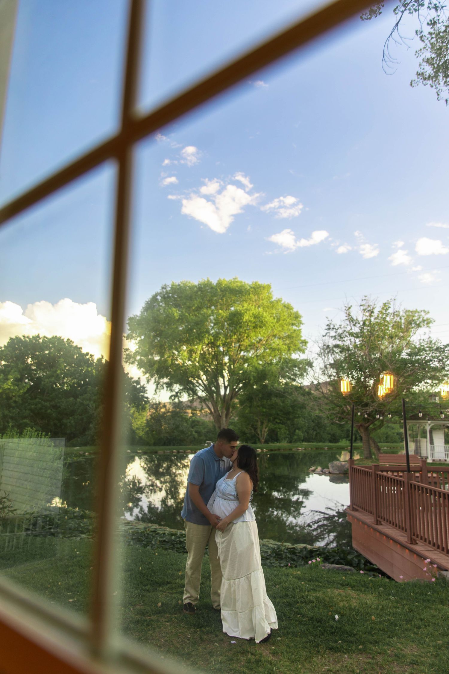 Romantic couple shares an intimate moment by a tranquil pond at sunset, viewed through a window pane.