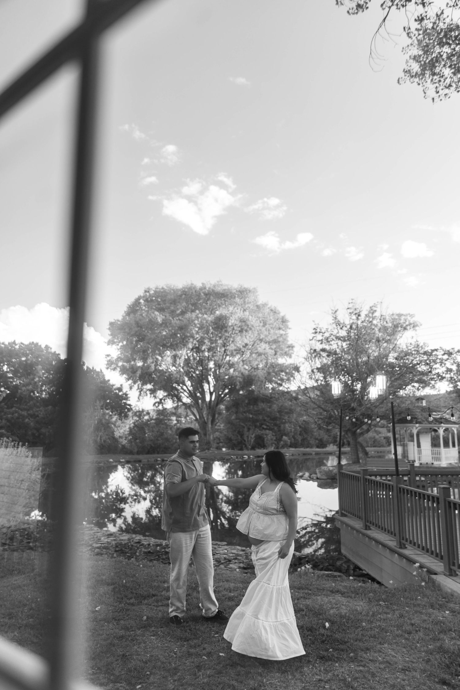 A black and white photograph of a couple walking on grass near a lake with trees in the background, framed through a window.
