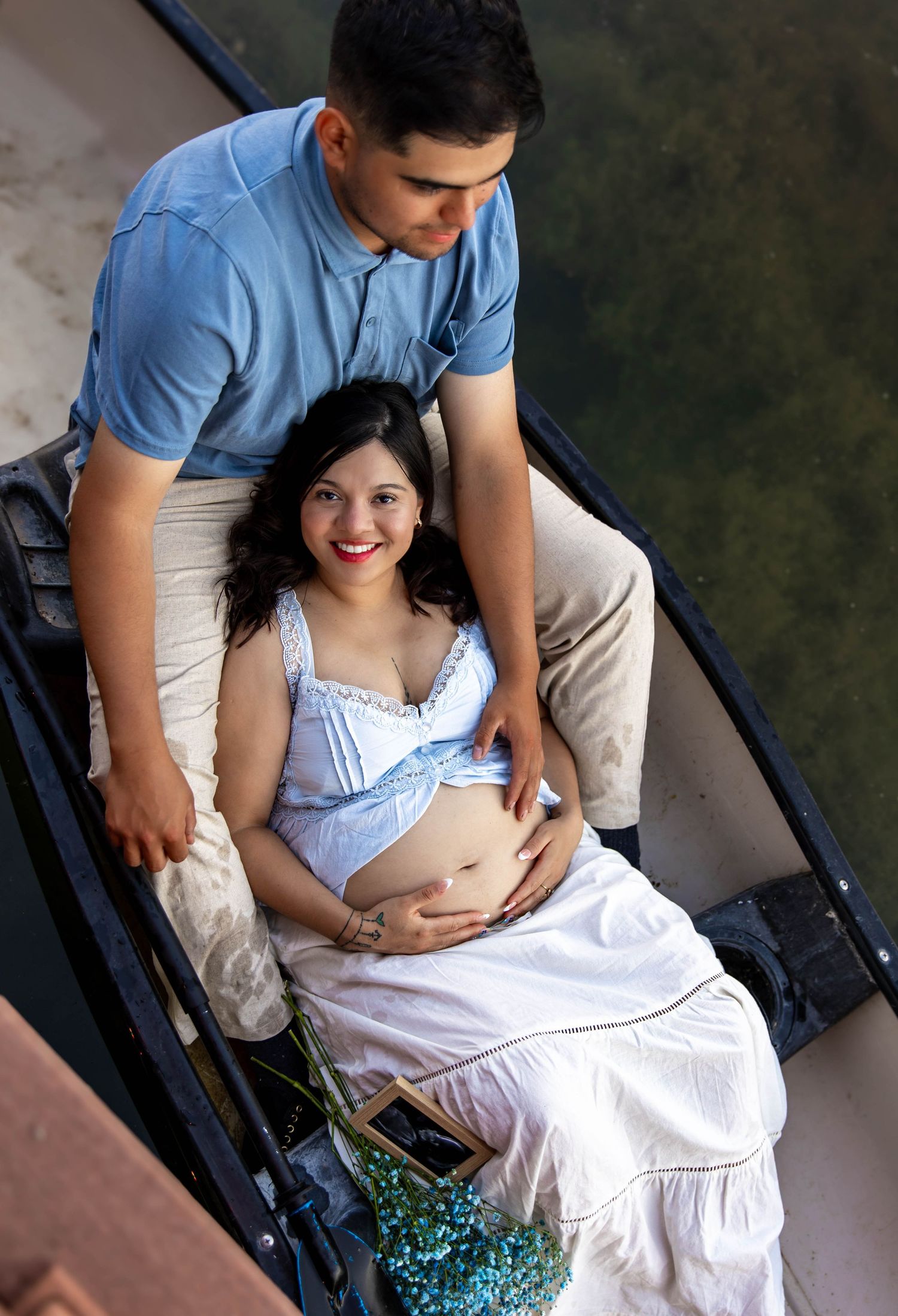 Pregnant woman in a white dress sits in a chair while her partner stands beside her in a tender outdoor setting.