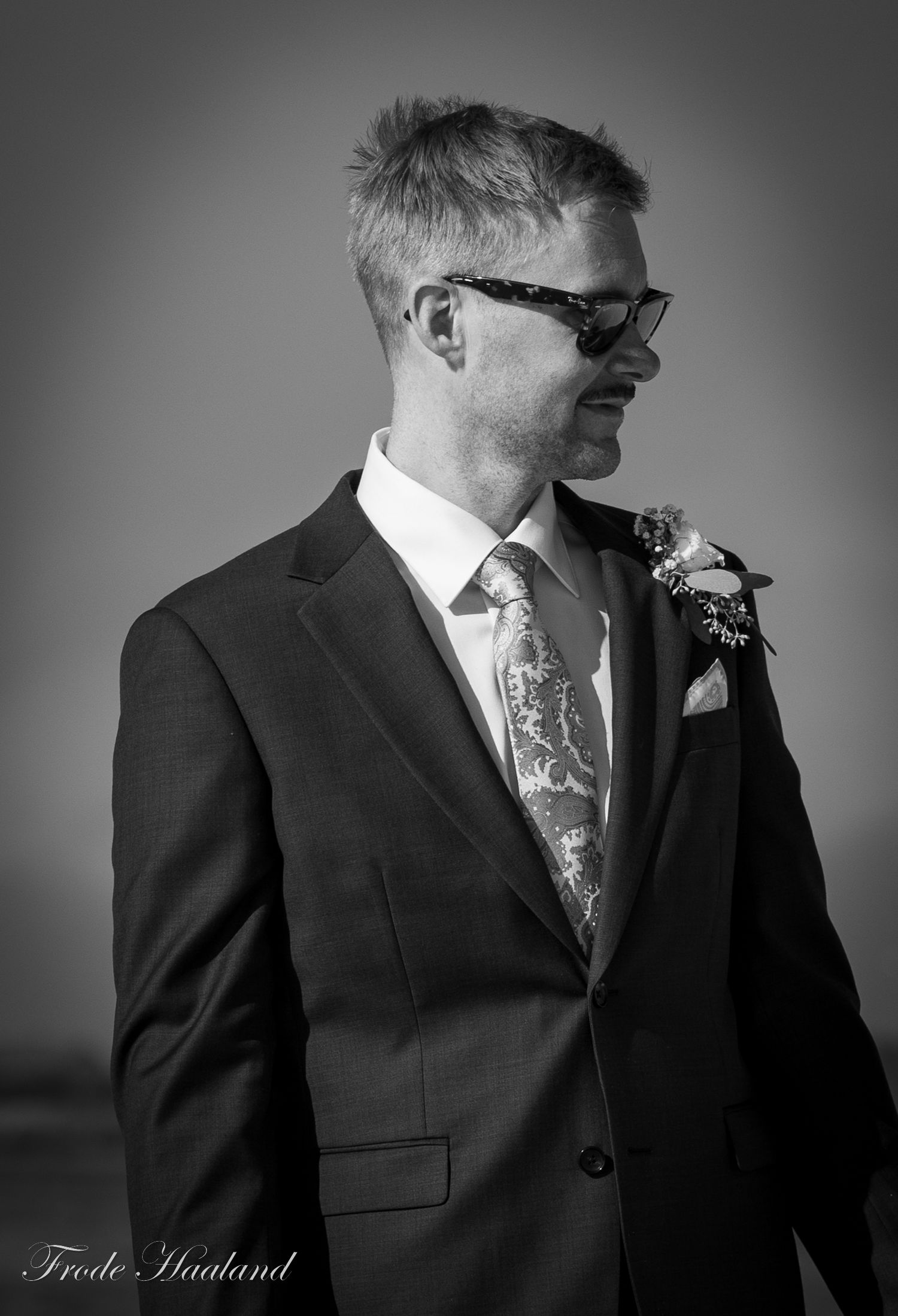 Stylish groom in dark suit with boutonniere and patterned tie in black and white portrait.