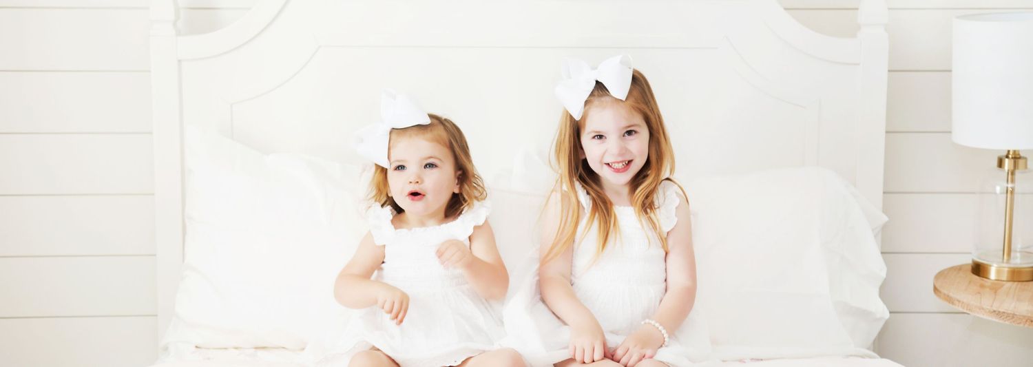 Two little kids in white dresses giggle together while sitting on a white bed.