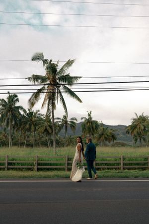 A couple walks down a road with palm trees and mountains in the Hawaiian landscape.
