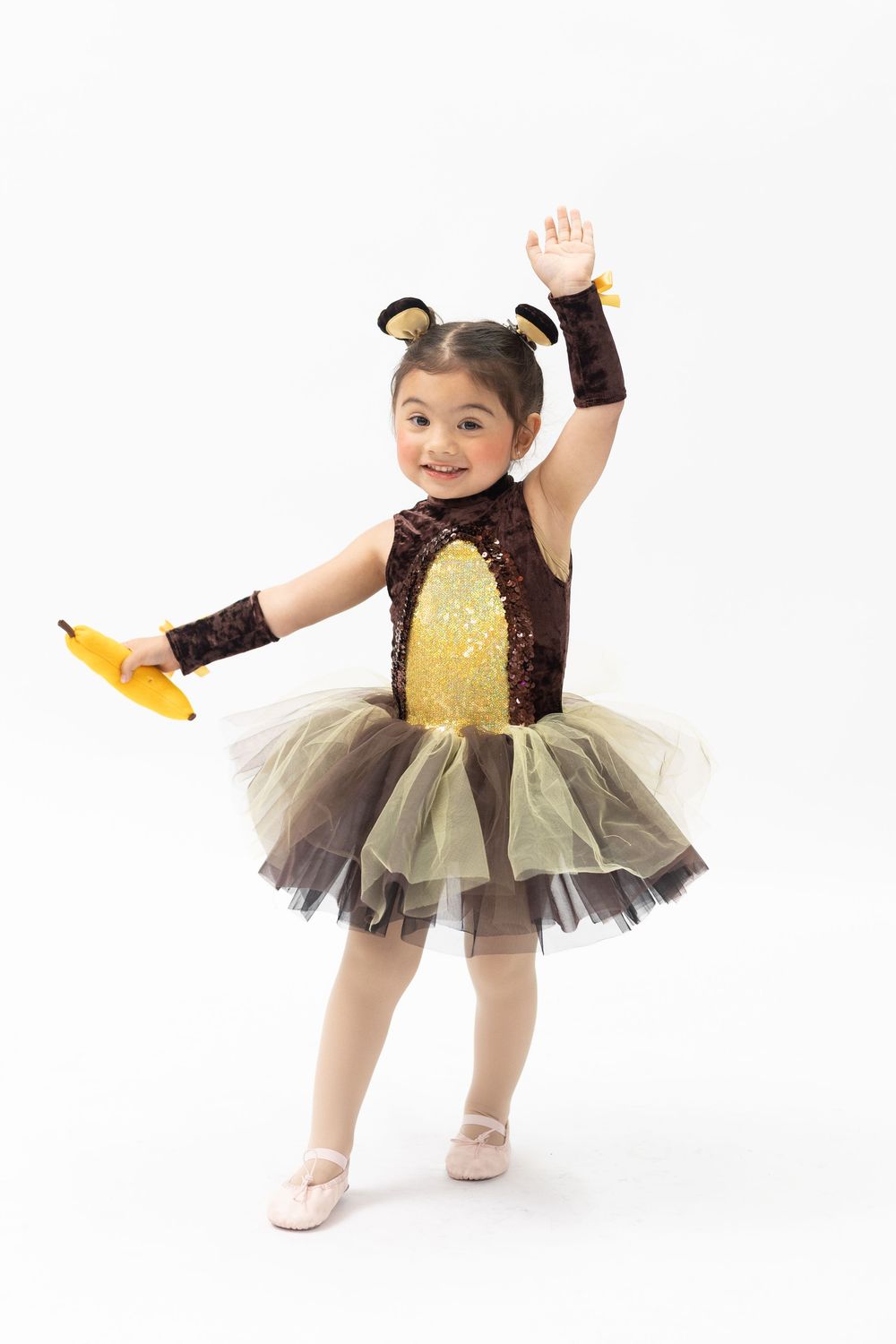 Young girl in monkey ballerina costume with tutu, holding banana, one arm raised, wearing ballet slippers on white background.