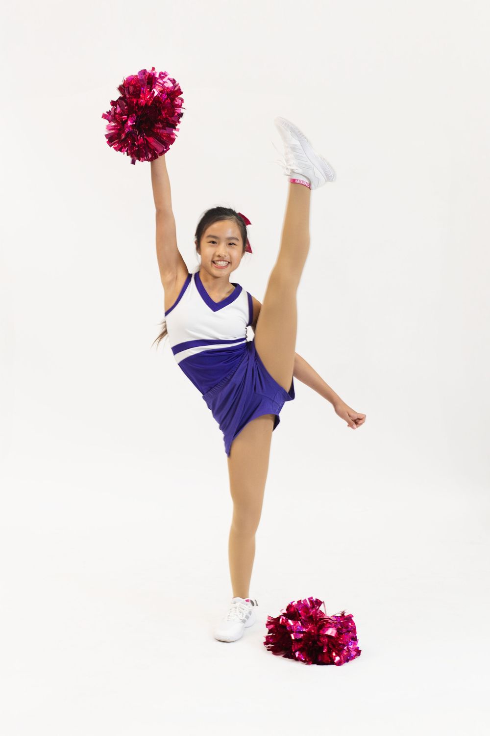 Young cheerleader in purple and white uniform performing high kick with red pom-poms on white background.