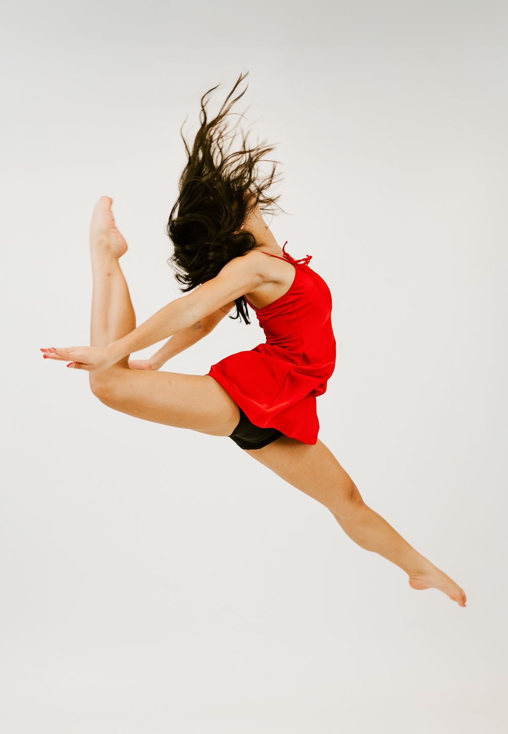 A female dancer in a red dress leaps gracefully mid-air, hair flowing, against a white background.