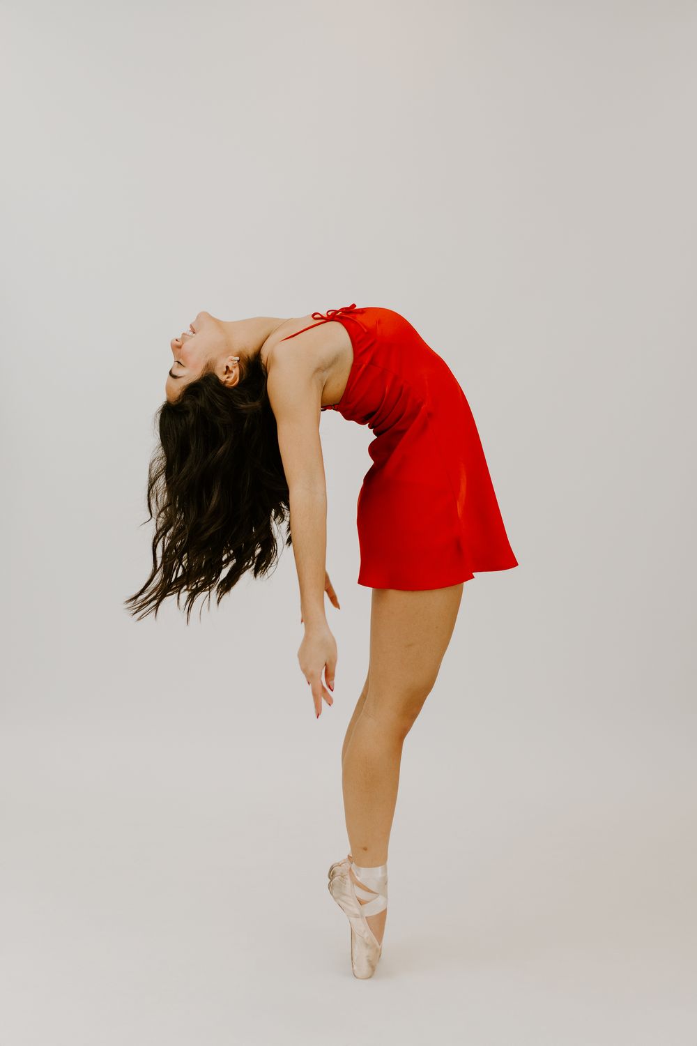 Graceful ballerina in red dress and pointe shoes performing an elegant backbend on white background.