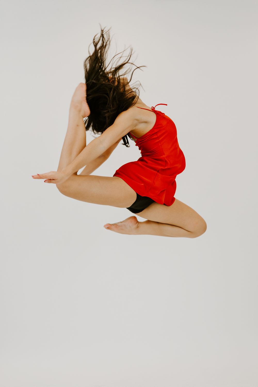 A dancer in a red dress leaps gracefully mid-air, hair flowing, against a white background.
