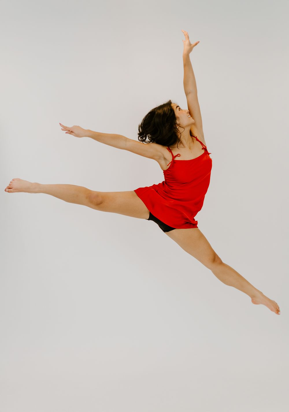 A female dancer in a red top leaps gracefully mid-air in a split jump against a white background.