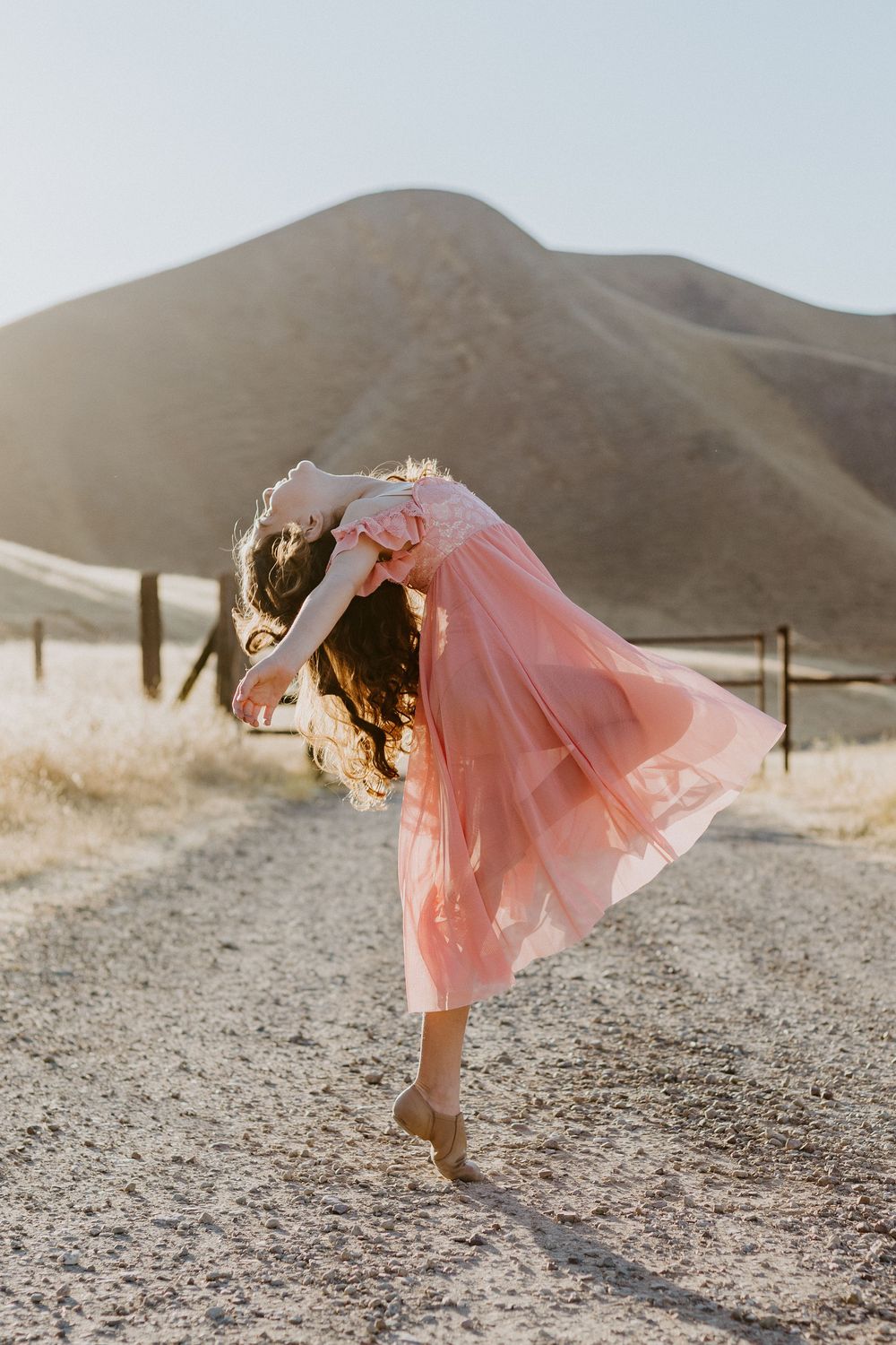 A girl in a pink tulle dress dances on a dirt path with golden hills in the background.