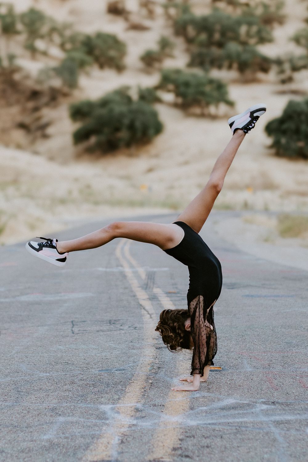 A gymnast performs a one-handed cartwheel on a desert road wearing a black leotard and sneakers.