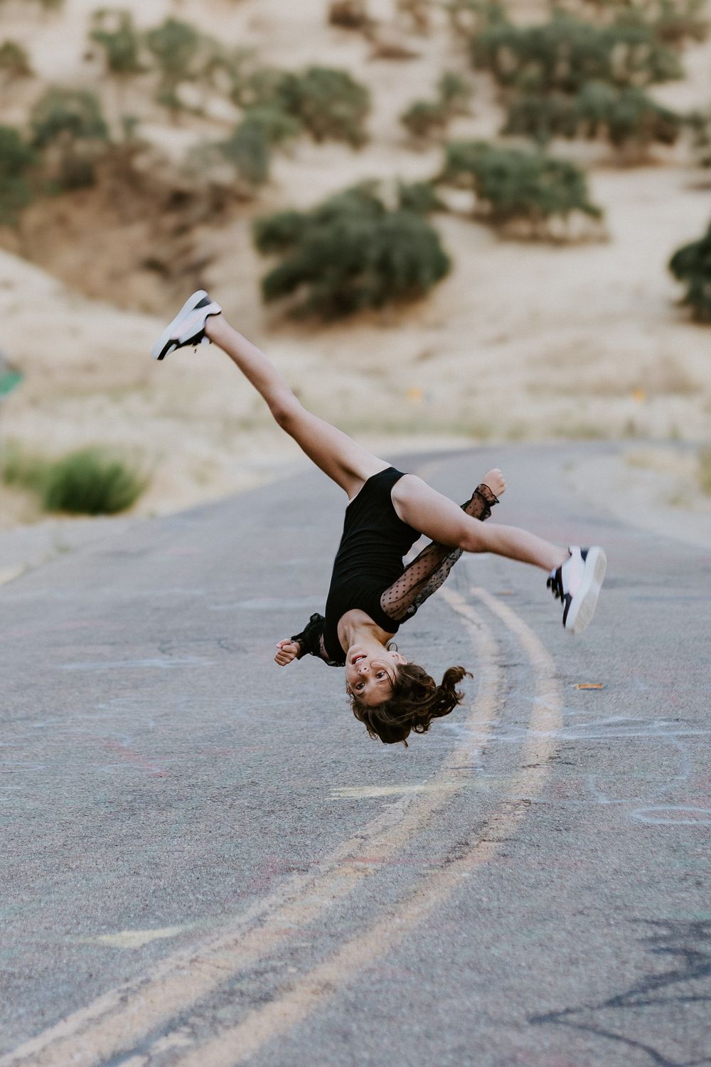 A child performs a backflip on a road in a desert landscape, wearing a black outfit and white sneakers.
