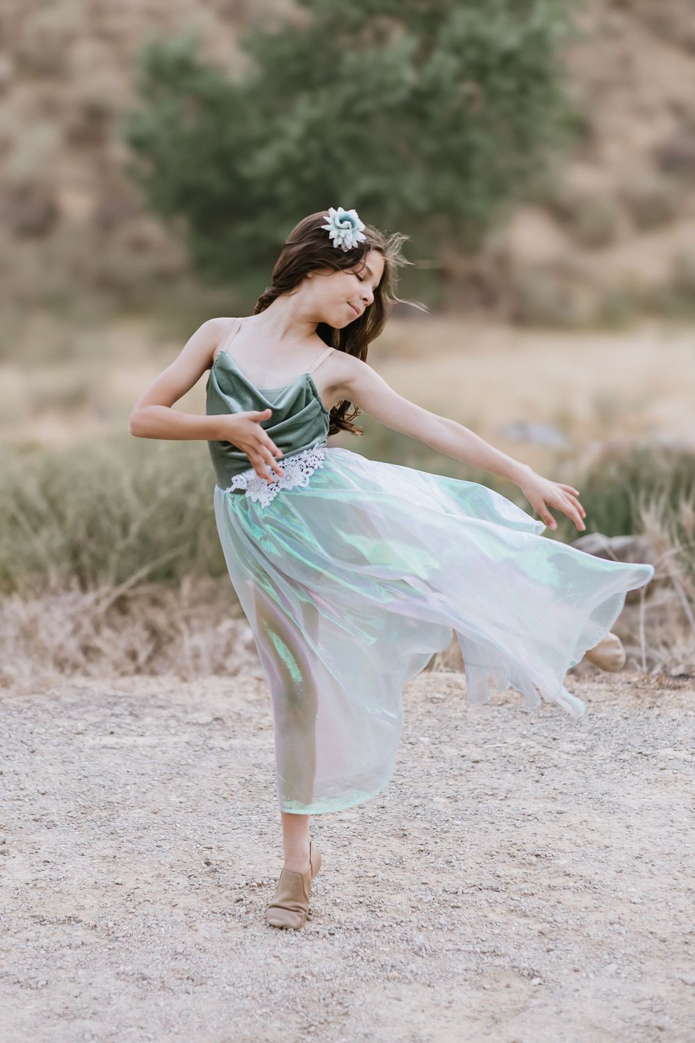 Young girl in flowing green tulle dress with floral crown dancing gracefully outdoors in a desert landscape.