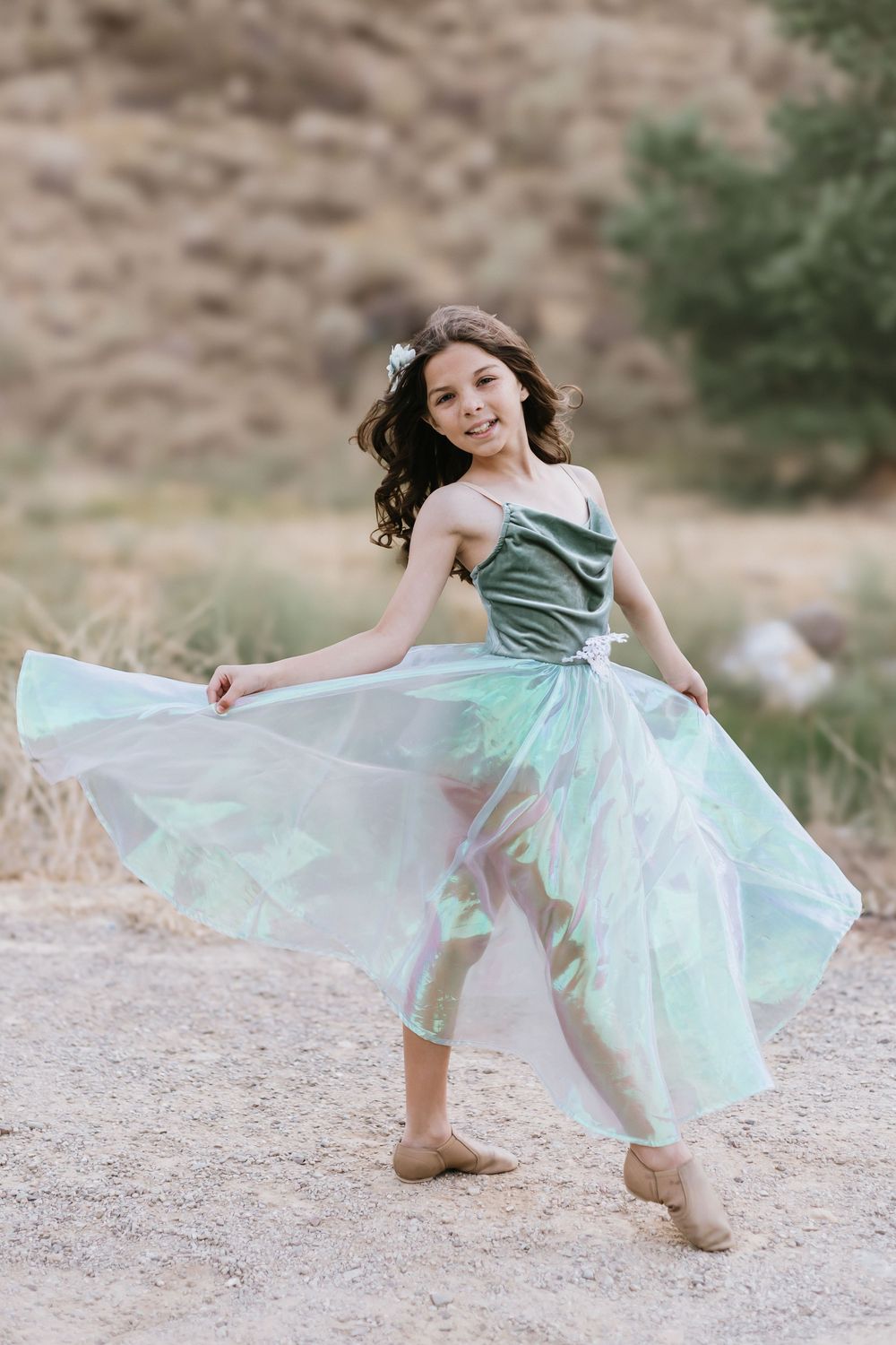 A young girl in a flowing iridescent green dress dances outdoors, smiling joyfully with rocky hills in the background.
