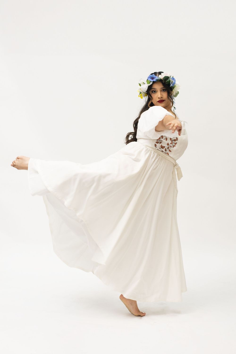 Woman in flowing white dress and floral crown dancing gracefully against white background.
