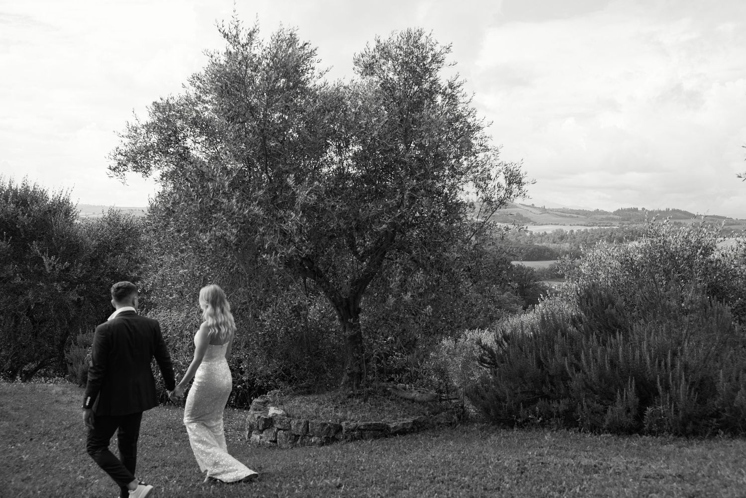 Black and white photo of a bride and groom walking hand in hand through a scenic garden with an olive tree.