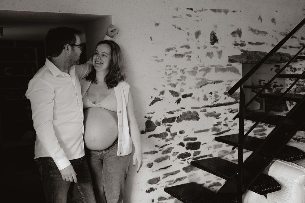 Black and white photo of a smiling pregnant couple standing together near a staircase and stone wall.