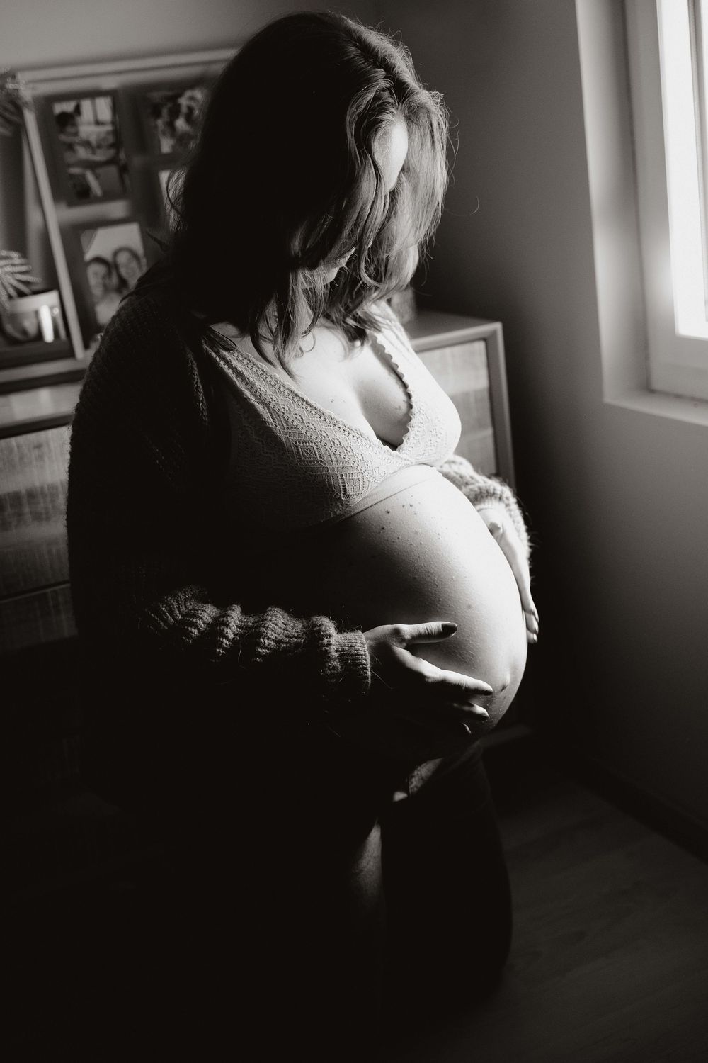 Black and white photo of a pregnant woman cradling her bare baby bump near a sunlit window.