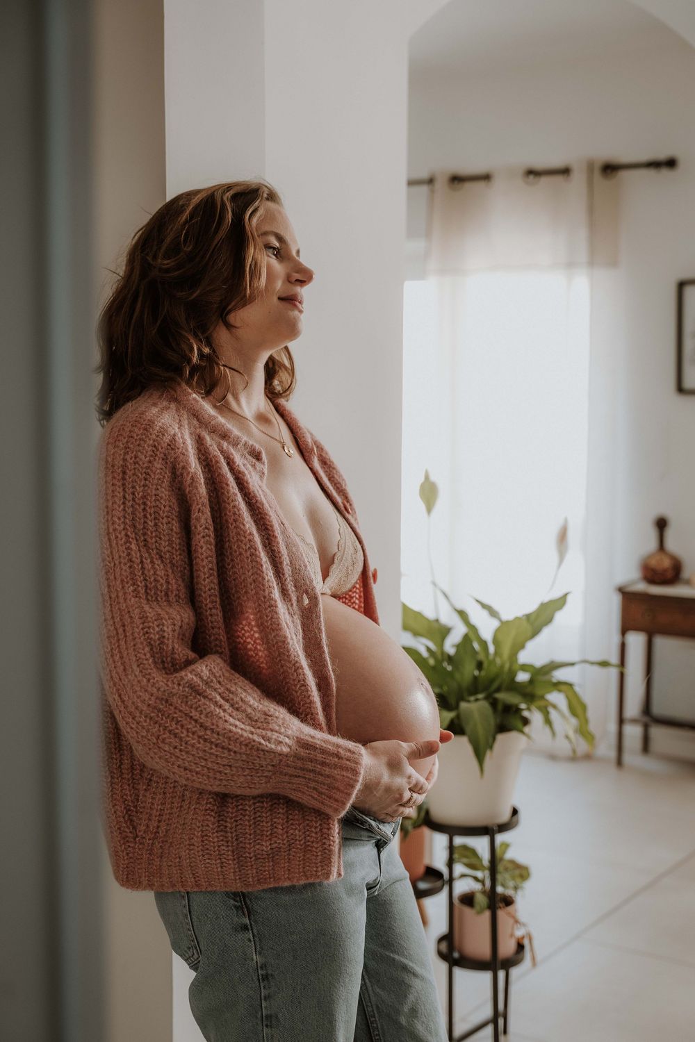 Pregnant woman in pink cardigan and jeans cradling her baby bump while standing by a window at home.