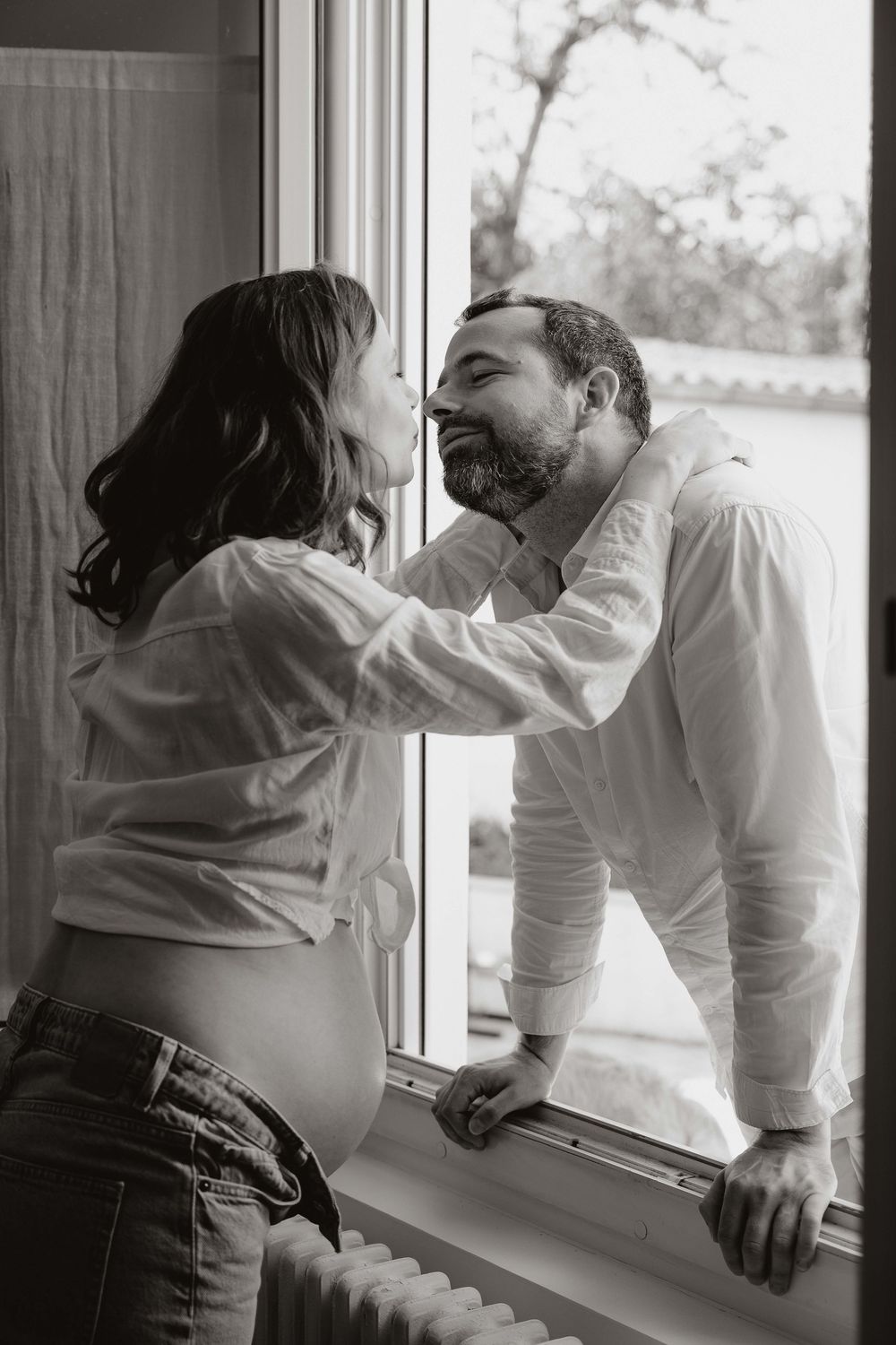 Black and white photo of a pregnant woman tenderly touching a bearded man's face by a window.