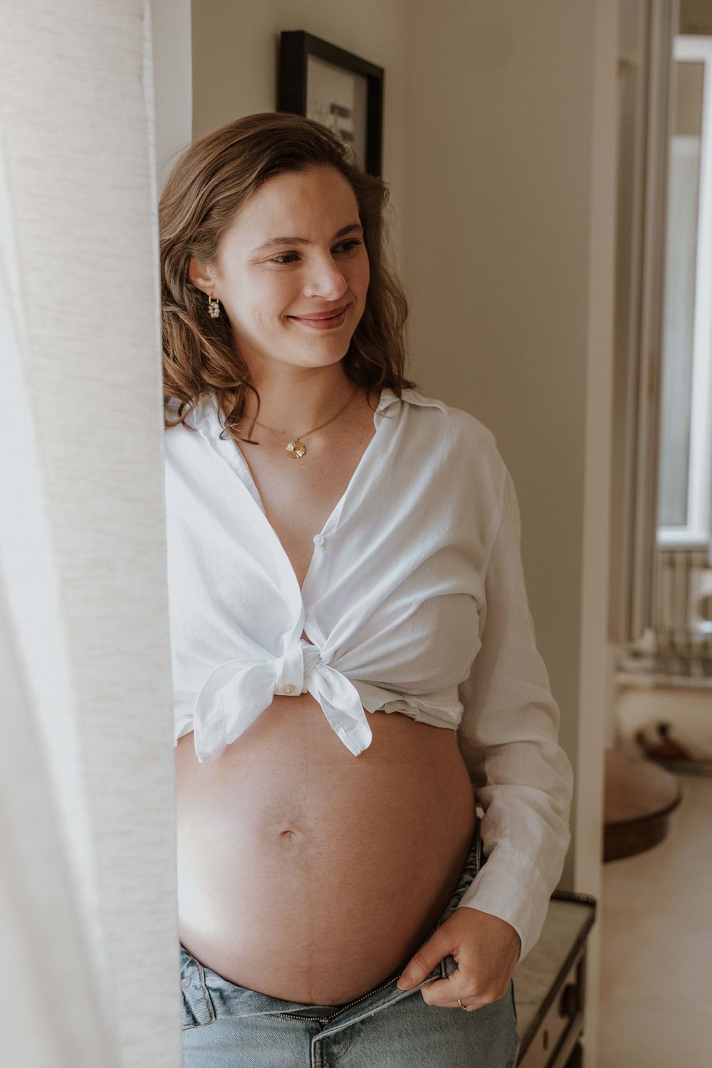 Smiling pregnant woman in white tied shirt and jeans showing baby bump near window.