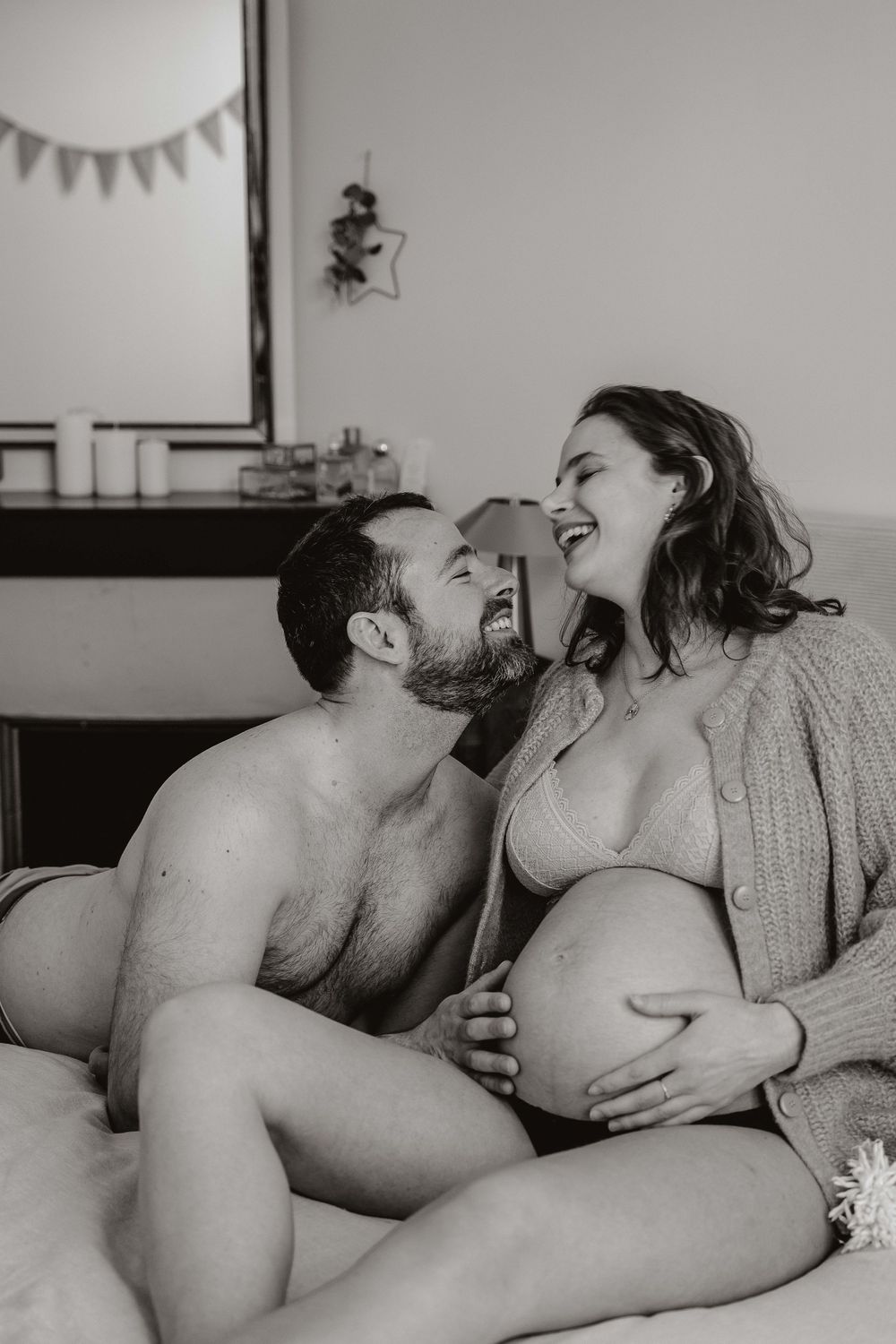 Black and white photo of a laughing pregnant couple sitting on a bed, sharing a joyful intimate moment.