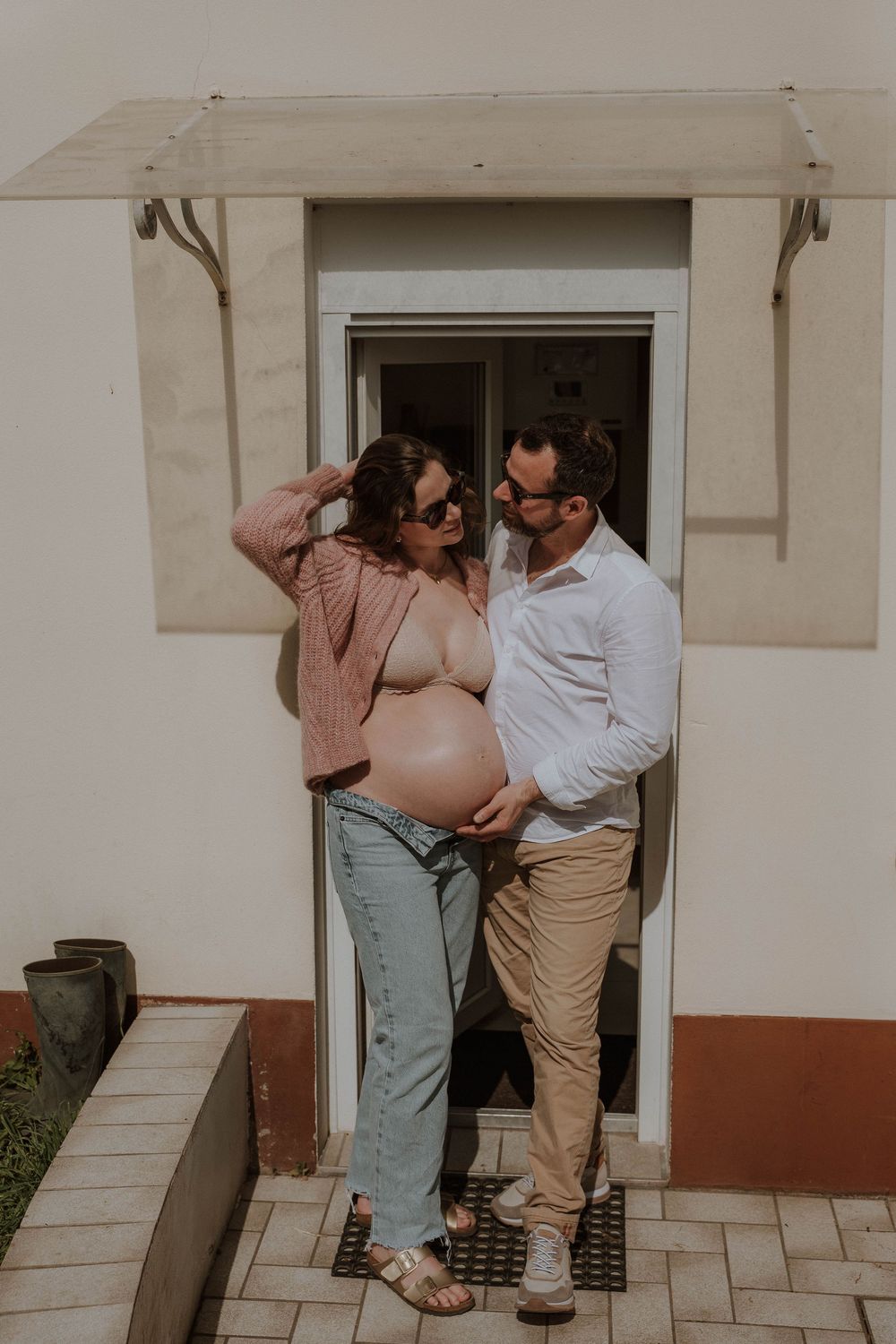 Couple posing outdoors, pregnant woman in pink cardigan and jeans with partner in white shirt, maternity photo shoot.