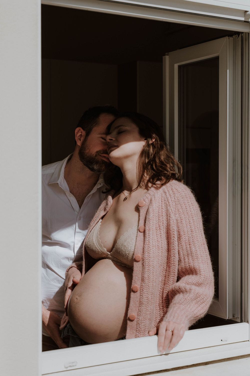 A couple shares a tender kiss by a sunlit window, the woman visibly pregnant in a pink cardigan and bikini top.