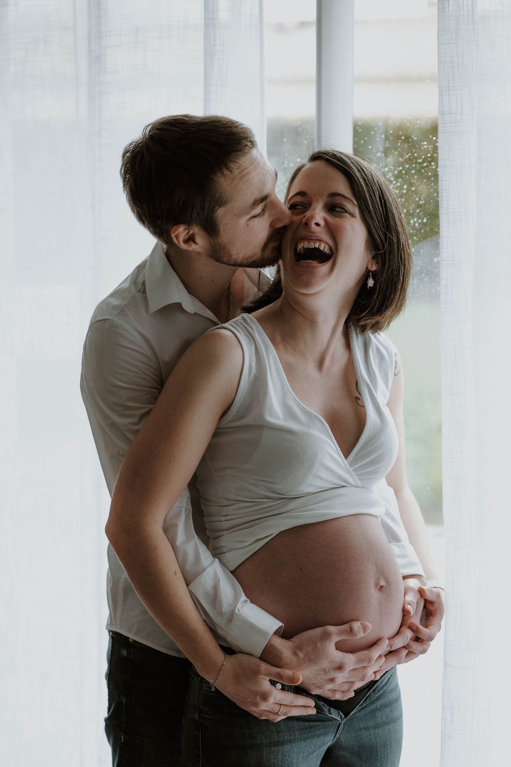 Happy pregnant woman laughing as her partner embraces her from behind, both cradling her baby bump near a bright window.