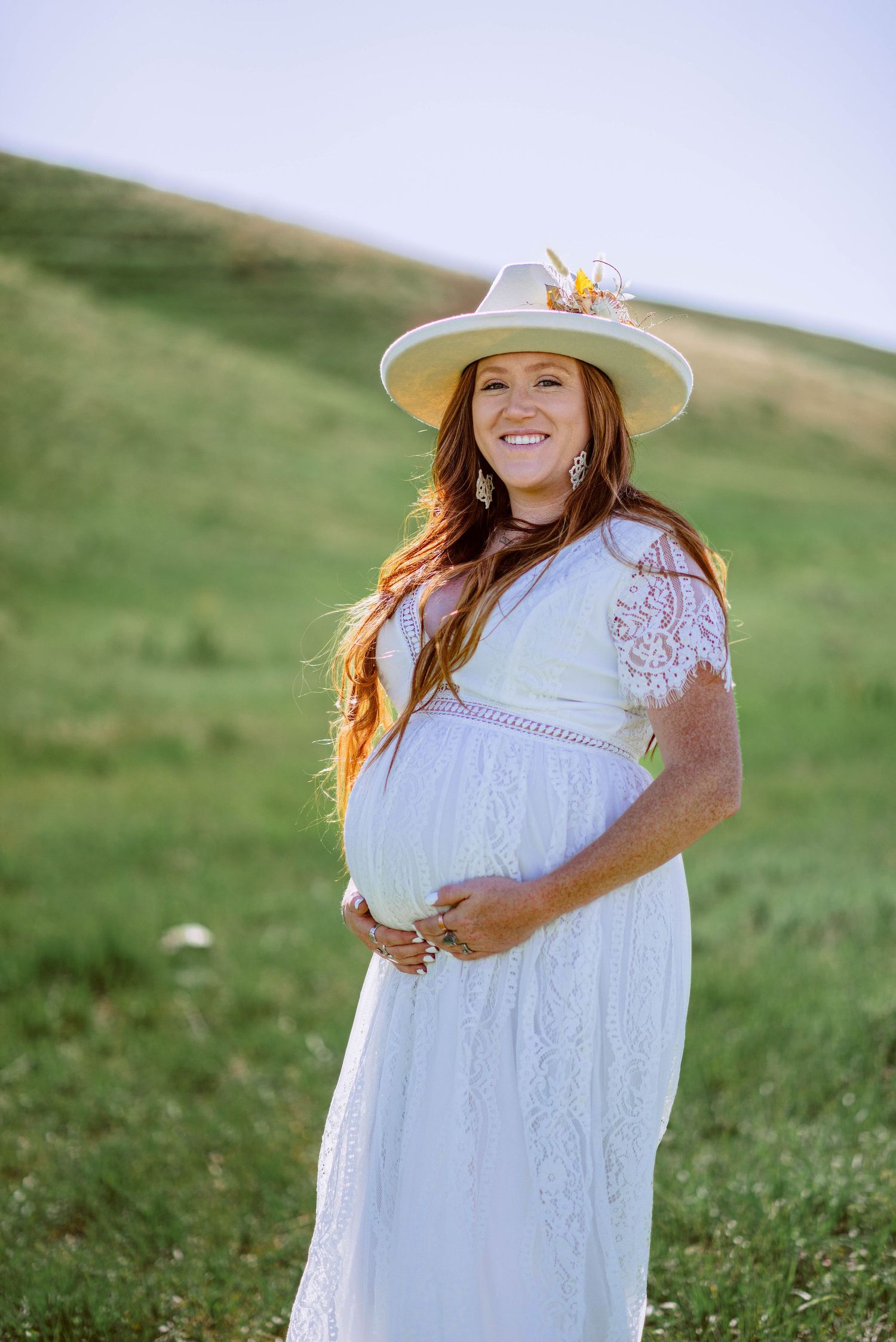 White lace maternity dress photoshoot in green rolling hills with decorative sun hat.
