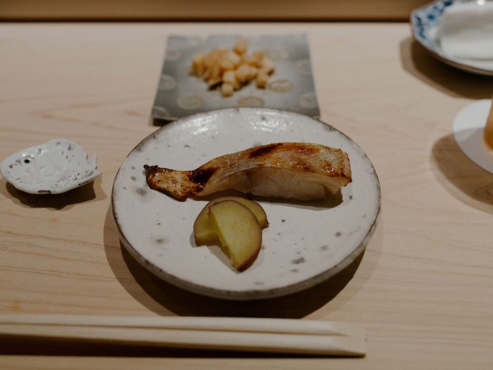 Grilled fish fillet served on a speckled ceramic plate with pickled ginger and garnishes.
