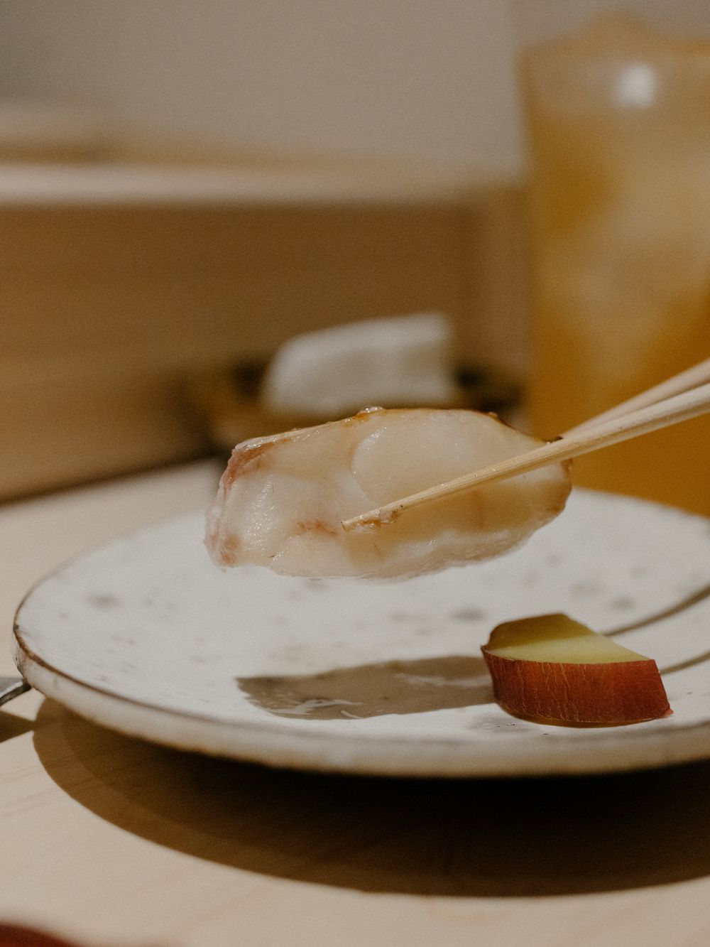 Delicate piece of white fish sashimi held by chopsticks over a plate with citrus garnish.