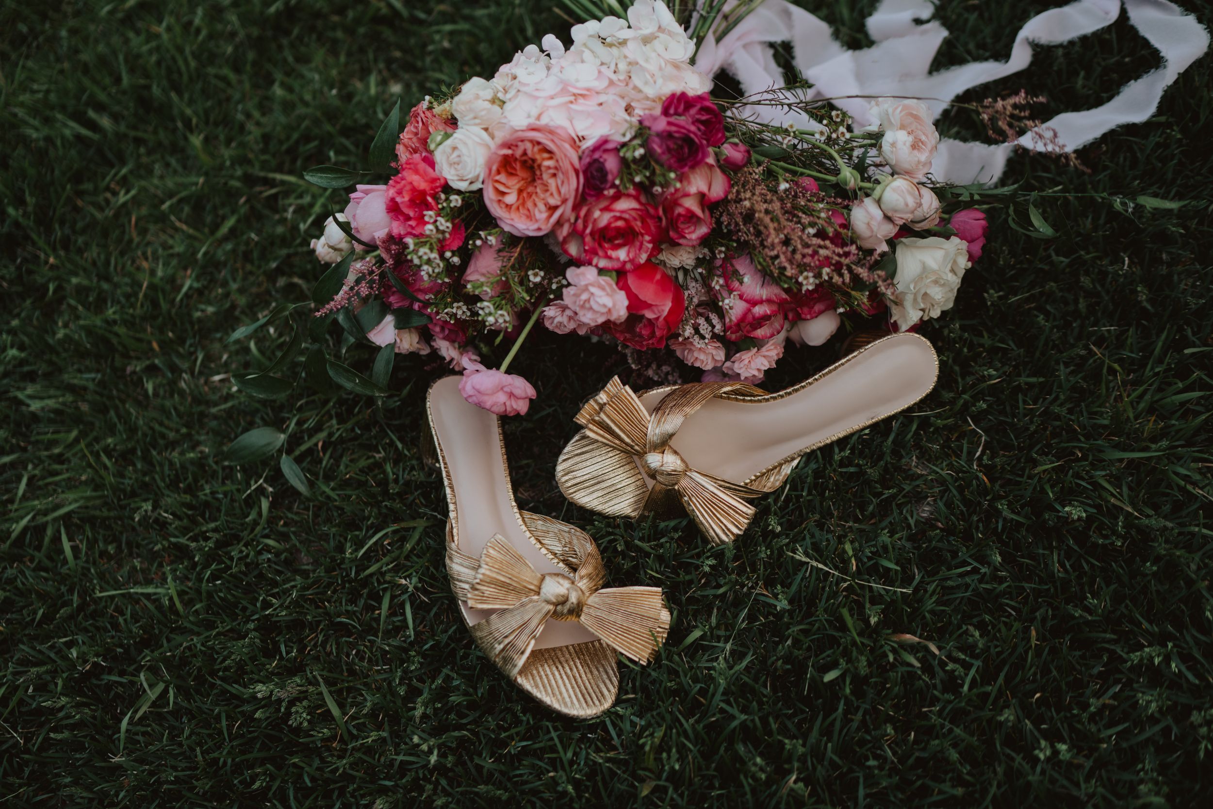Pink and red wedding bouquet with elegant gold bow sandals on grass.