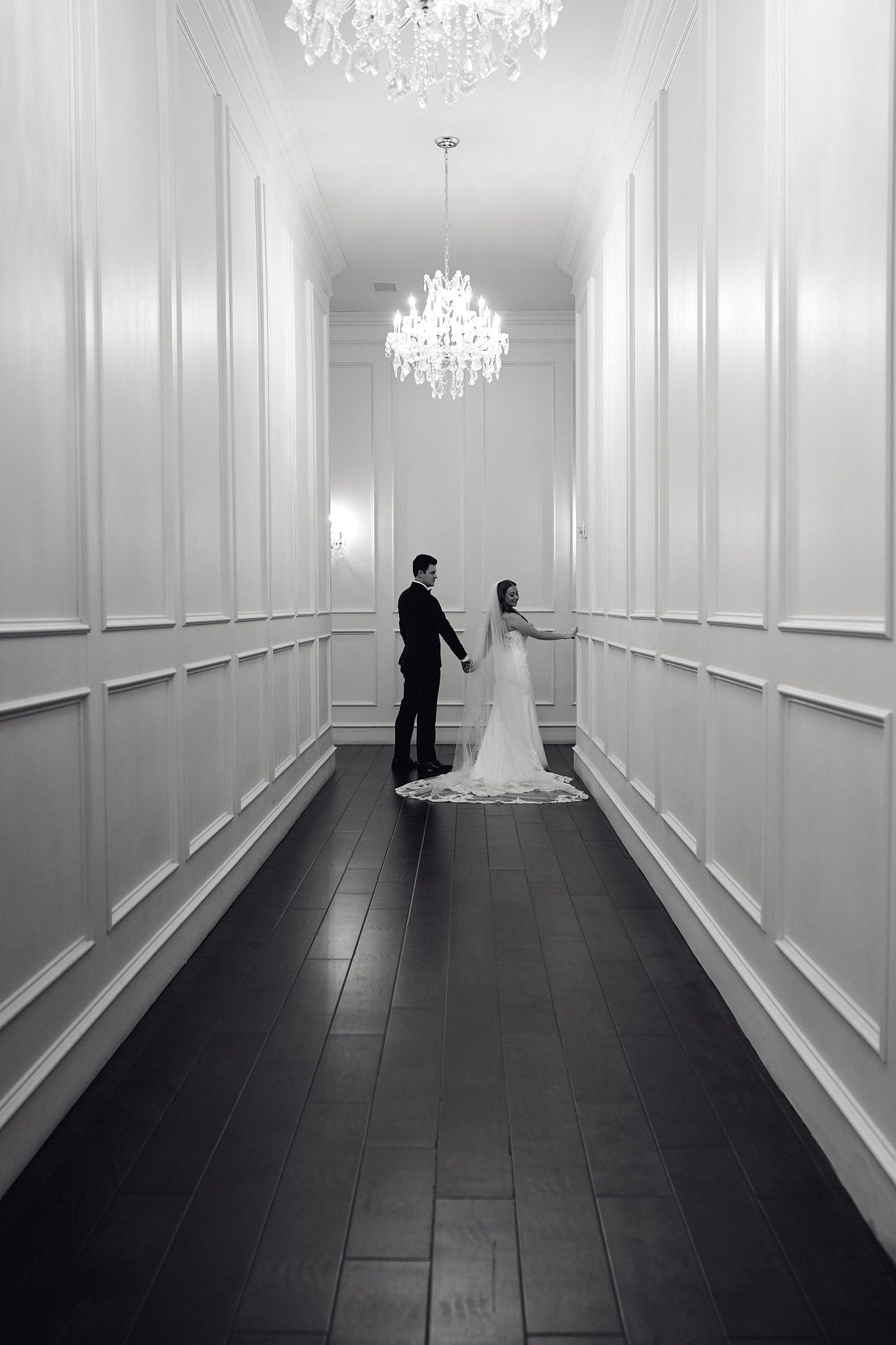 Black and white photo of a couple in formal wear standing in an elegant hallway with crystal chandelier.