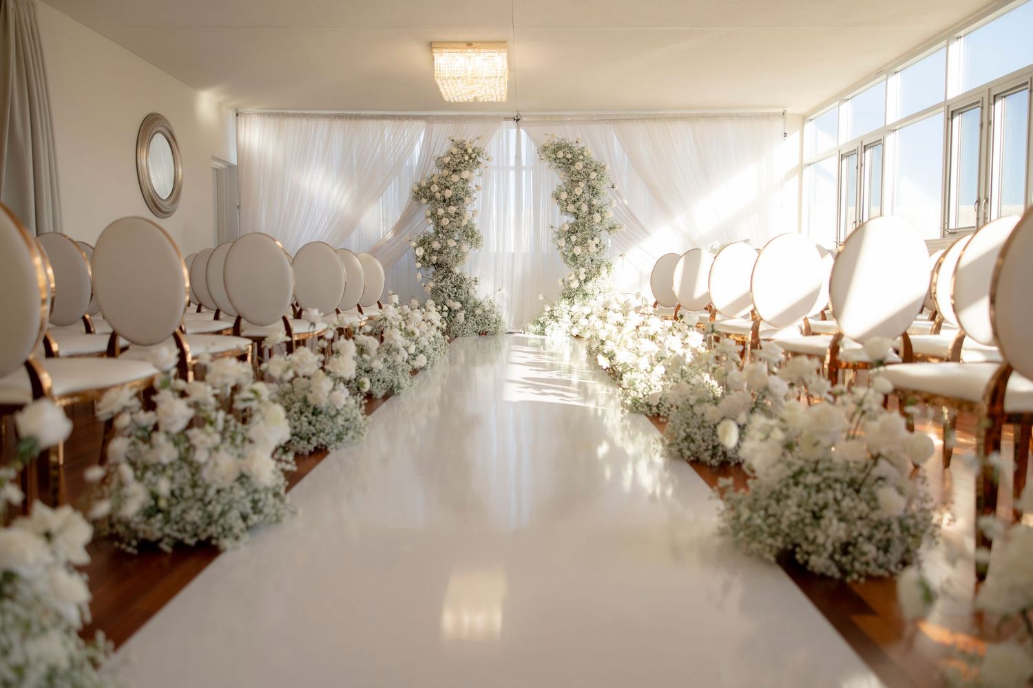 Elegant white wedding ceremony setup with floral aisle decorations and white chairs in sunlit room.