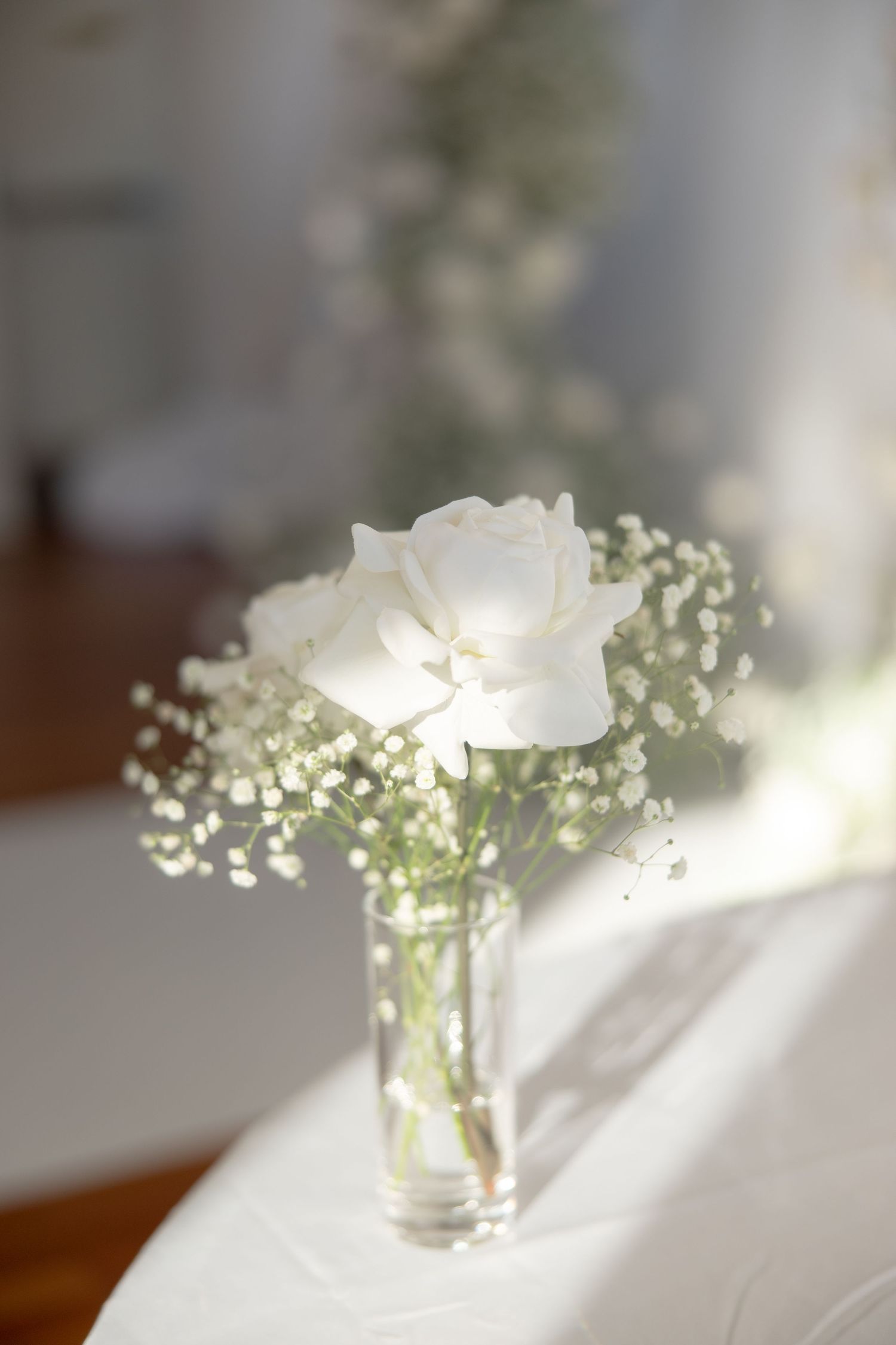 Single white rose and baby's breath in glass vase on white tablecloth.