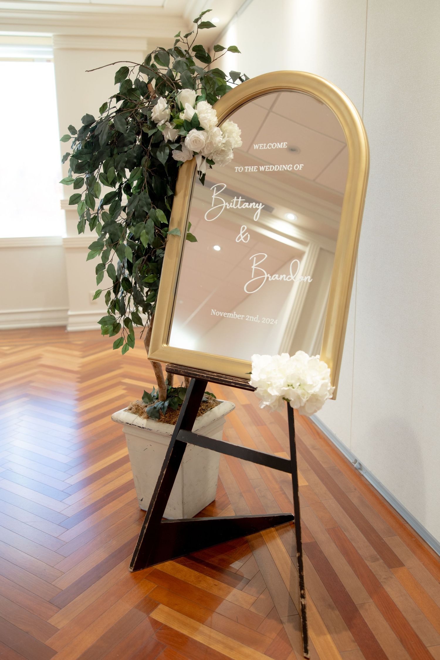 Gold-framed wedding welcome sign decorated with white roses and greenery on wooden floor.
